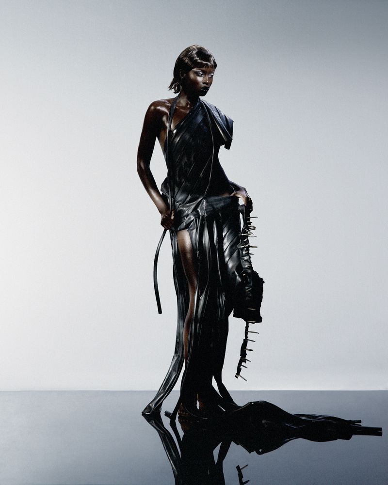 Woman in avant-garde black dress with spike accents, posing against a white studio backdrop with reflective floor, showcasing high fashion.
