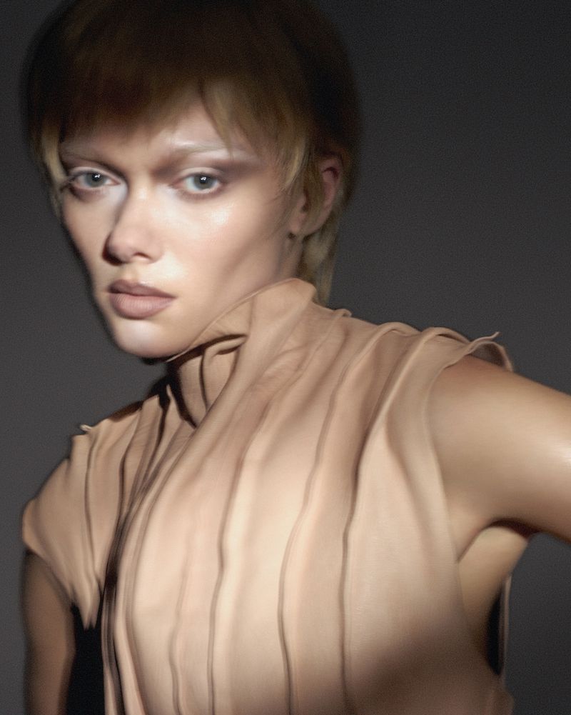 Woman in beige cowl-neck top with short blonde hair, posing indoors in studio lighting against a dark gray backdrop for a fashion shot.
