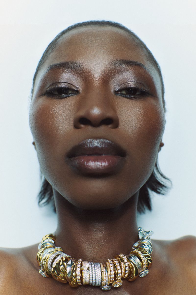 Close-up portrait of a dark-skinned woman wearing an elaborate gold and silver ring necklace against a plain white background.