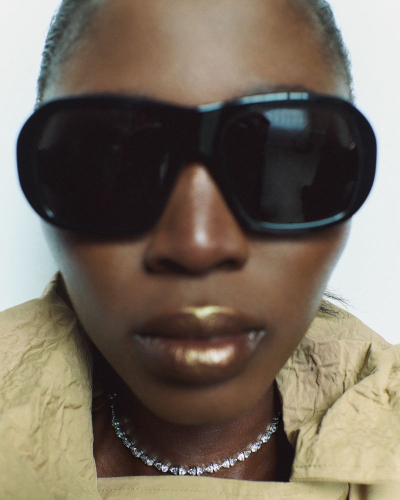 Close-up of a Black woman with sunglasses and gold lipstick, wearing a diamond necklace against a bright white backdrop in soft lighting.