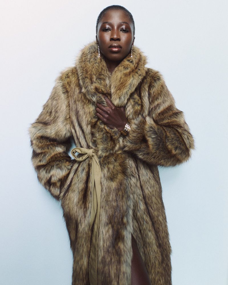 Elegant Black woman wearing a fur coat, posed in front of a simple white wall, showcasing fashion, style, and beauty.