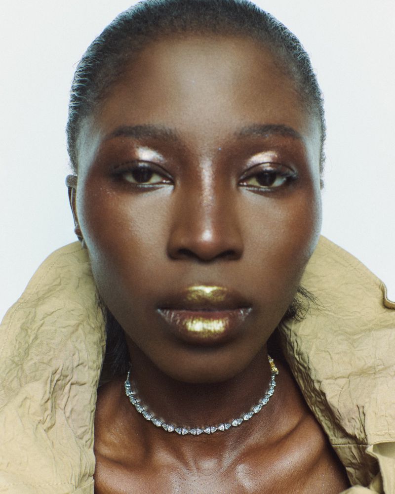 Close-up portrait of a Black woman with gold lipstick and a clear jeweled necklace against a white background, wearing a beige jacket.