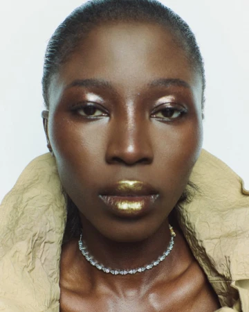 Close-up portrait of model Haddy Ceesay wearing metallic gold lipstick and a diamond choker collar against a white backdrop.