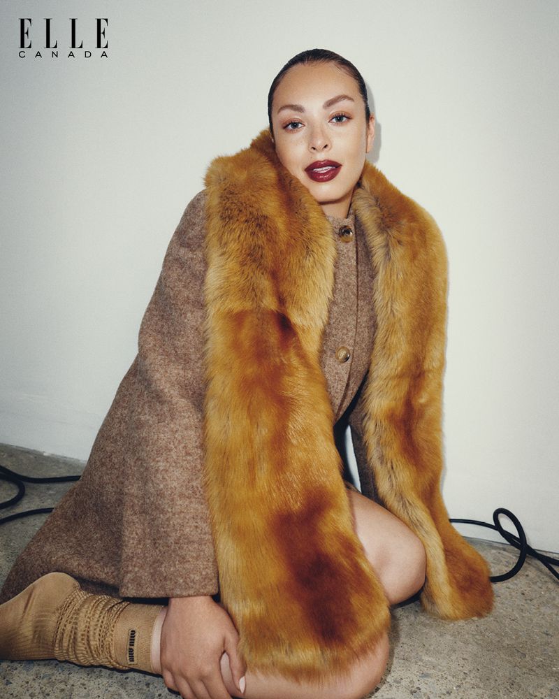 Woman in brown coat with fur collar poses kneeling indoors for a fashion shot with Elle Canada magazine logo visible in corner.