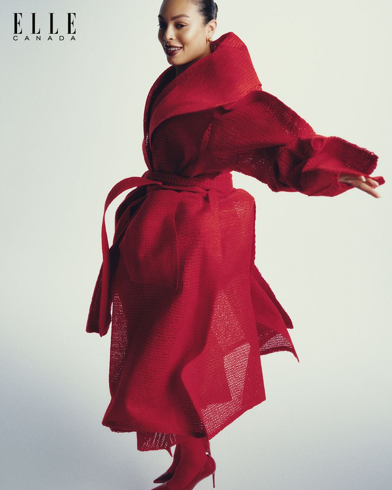 Editorial fashion shot of an adult woman in a red textured coat with a belt, and red heels, posing against a white background.