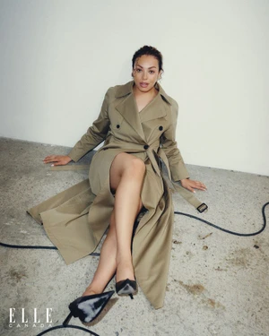 Sarah Nurse seated on a concrete studio floor wearing a belted beige trench coat and black pointed heels for Elle Canada.
