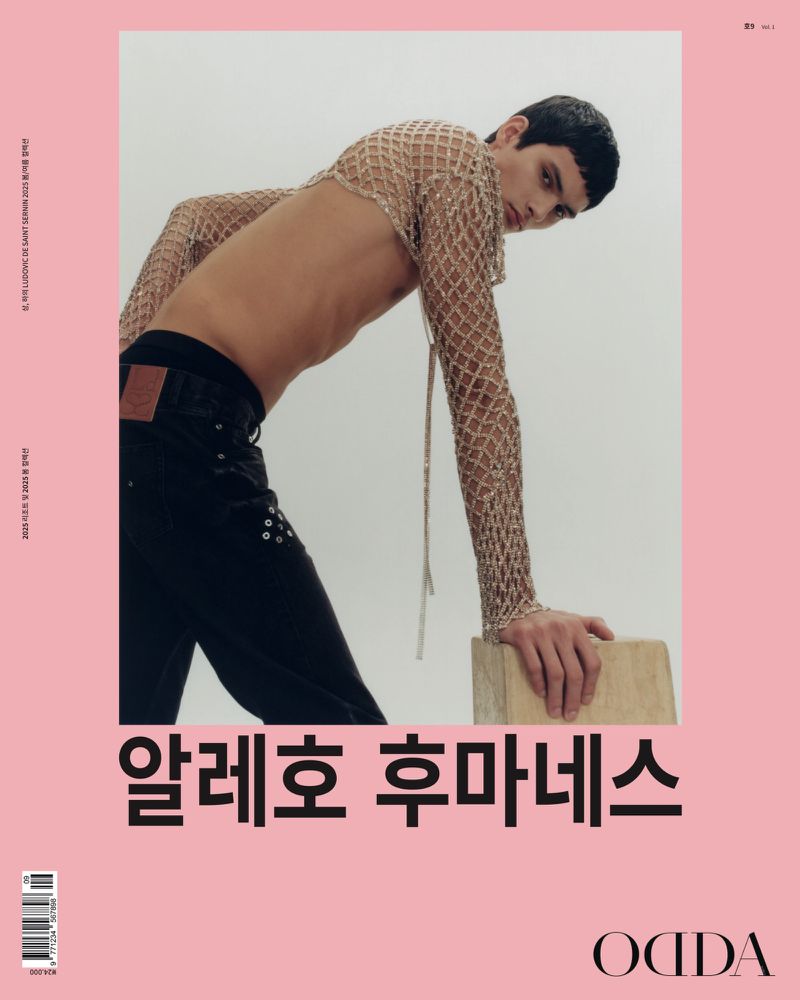 A fashion editorial magazine cover shot featuring a male model in a net shirt leaning on a wood block against a light pink background.