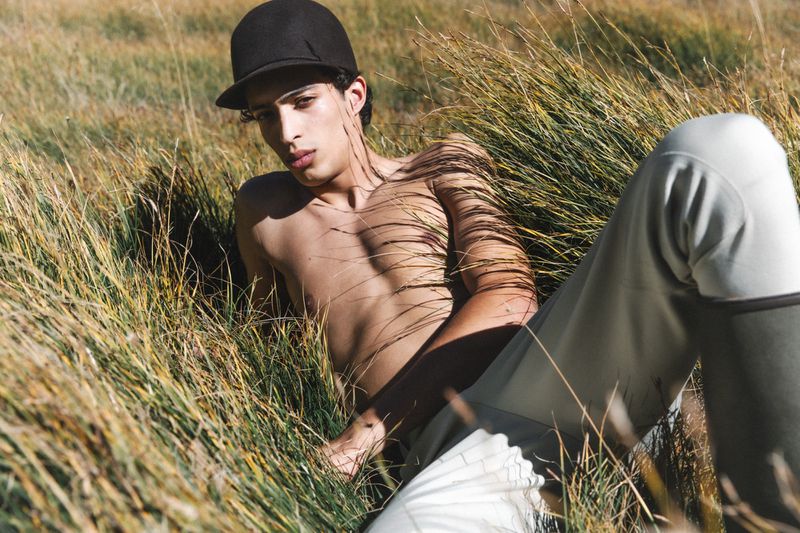 Shirtless adult male with black cap lies in a grassy field outdoors in the daytime, wearing light-colored pants, looking at the camera.