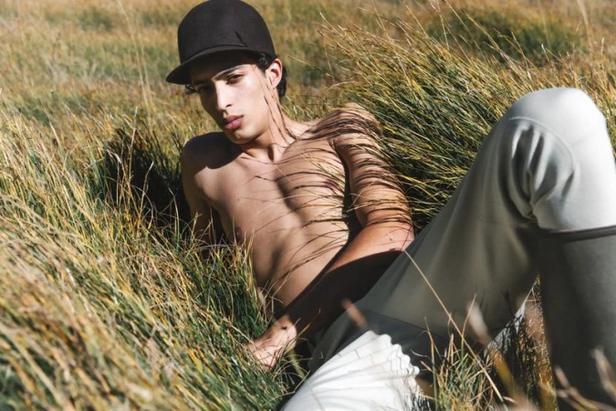 Shirtless adult male with black cap lies in a grassy field outdoors in the daytime, wearing light-colored pants, looking at the camera.