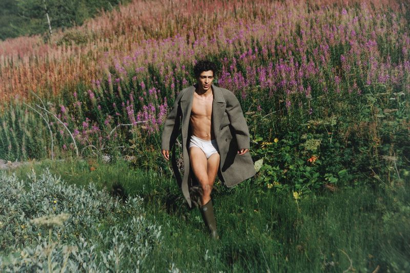 A man in white underwear and a coat walks through a field of purple flowers and tall grass on a sunny day in a rural setting.