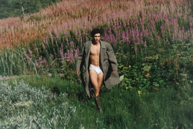 A man in white underwear and a coat walks through a field of purple flowers and tall grass on a sunny day in a rural setting.