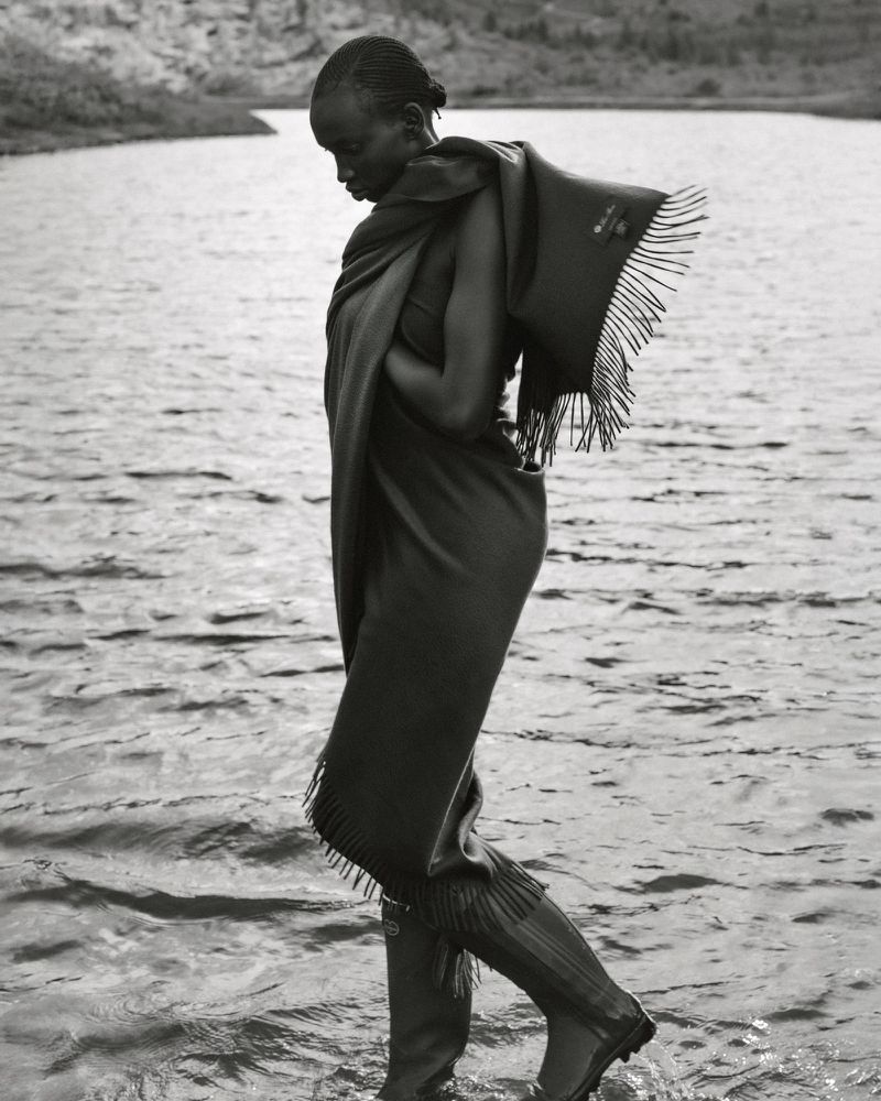 Black and white portrait of an African woman in a wrap and boots wading through a lake with hills visible in the background.