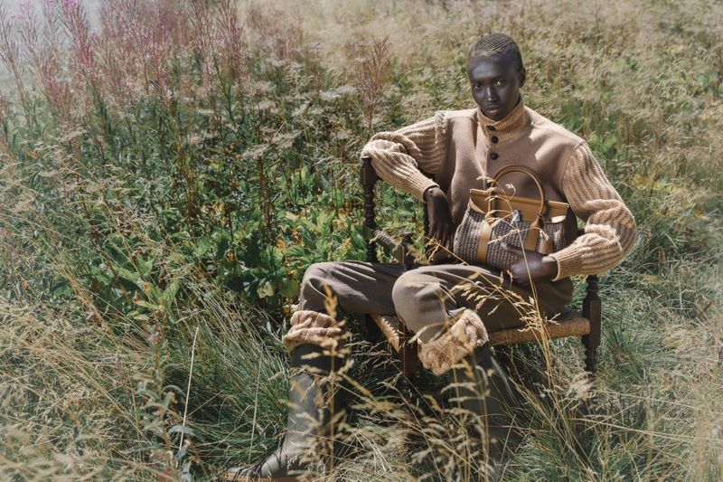 An adult poses seated in a chair outdoors in a field of tall grasses, wearing a knitted sweater and holding a handbag in a fashion editorial.