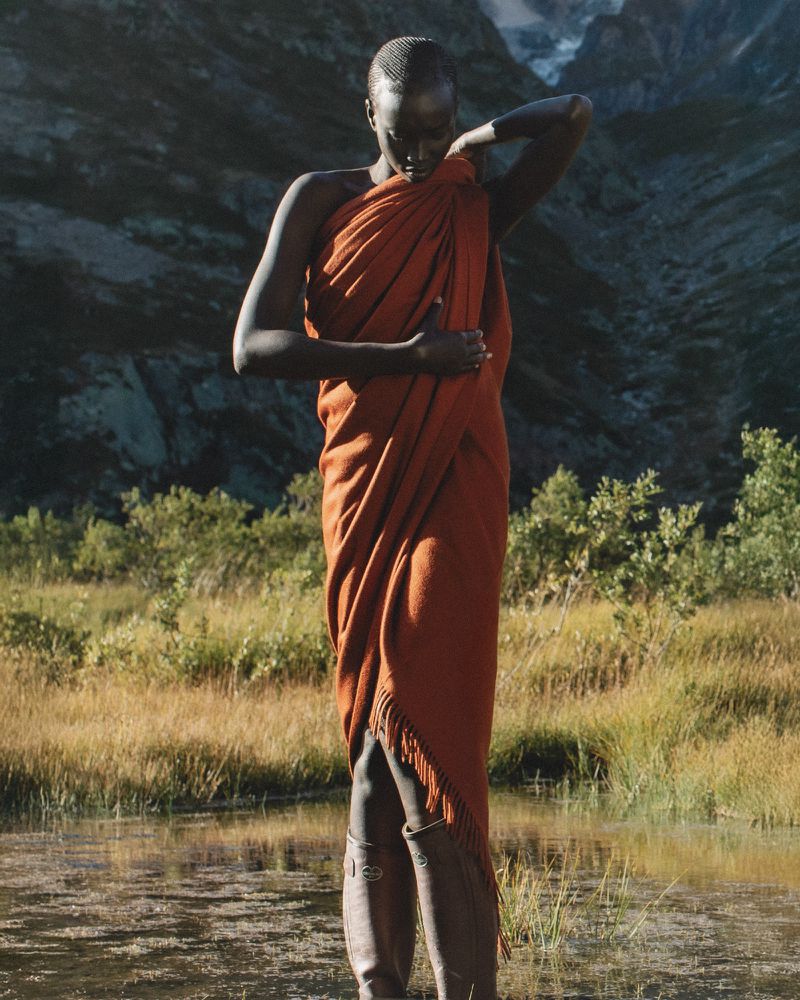 Woman draped in orange fabric standing in a body of water surrounded by grassy land and mountains in the background on a sunny day.