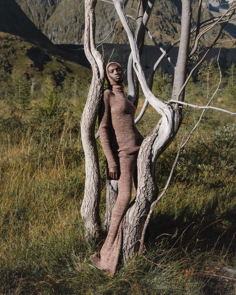 An adult female fashion model poses in a knit dress, leaning against a light-colored tree in a grassy meadow with a mountain backdrop.