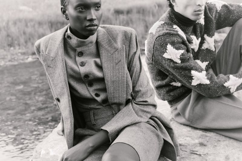 Monochrome fashion shot of a black woman in a grey suit with a man in a star-patterned sweater sitting on a rock in a natural setting.