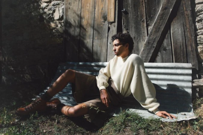 Young adult male poses in off-white sweater and brown shorts, lounging on corrugated metal with grass in front of a weathered wooden building.