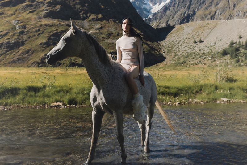 An adult female riding a grey horse through shallow water in a mountain landscape with snowy peaks and green hills, daytime.