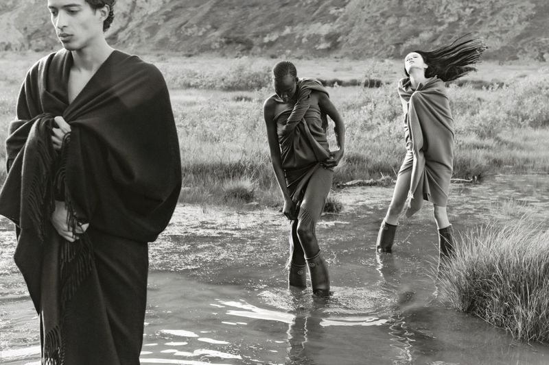 Three models pose in ankle-deep water with a mountain backdrop. They are draped in flowing fabrics, creating a monochrome fashion editorial.