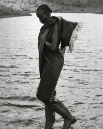 Black and white fashion photograph of a woman wrapped in a fringed blanket wading through water in knee-high rubber boots.