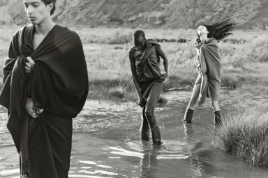 Three models wearing draped shawls standing in shallow water outdoors with rugged terrain in the background