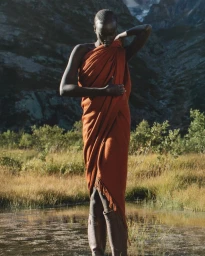 Model draped in a burnt orange blanket standing in a shallow river with a mountain backdrop