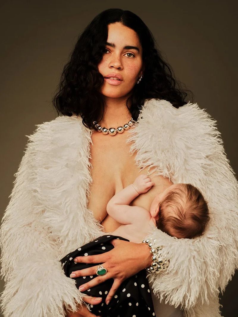 Adult female breastfeeding a baby, wearing a fluffy white jacket, jewelry, and a polka dot skirt against a grey background.