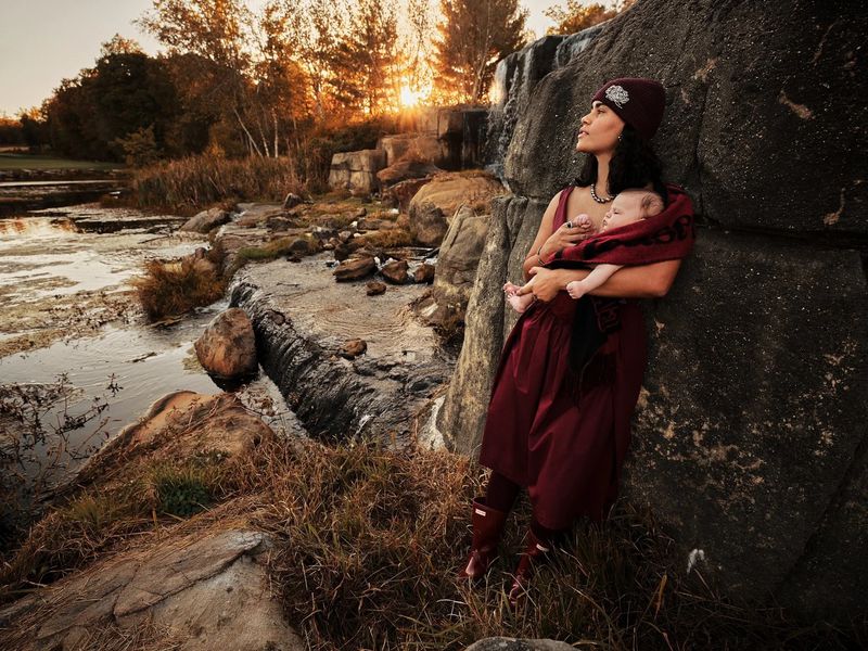 Mother holds her infant child near a rock face at golden hour. She wears a burgundy dress and hat in a nature setting.
