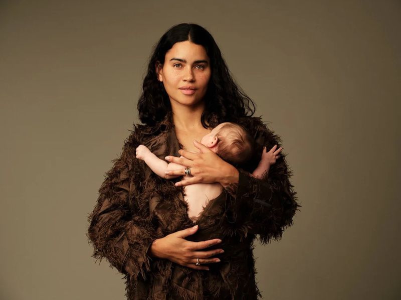 Portrait of a woman in a textured coat holding a baby against a grey background. The woman is gazing toward the camera.