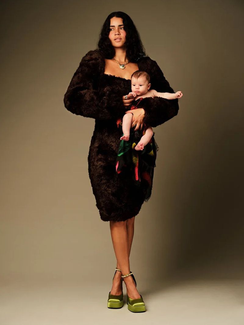 A stylish studio shot of a woman in a brown textured dress and green platform heels, holding a baby in a colorful garment.