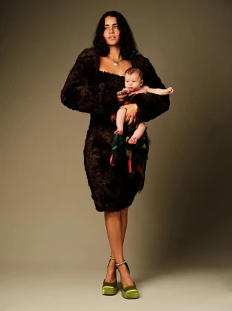 A stylish studio shot of a woman in a brown textured dress and green platform heels, holding a baby in a colorful garment.