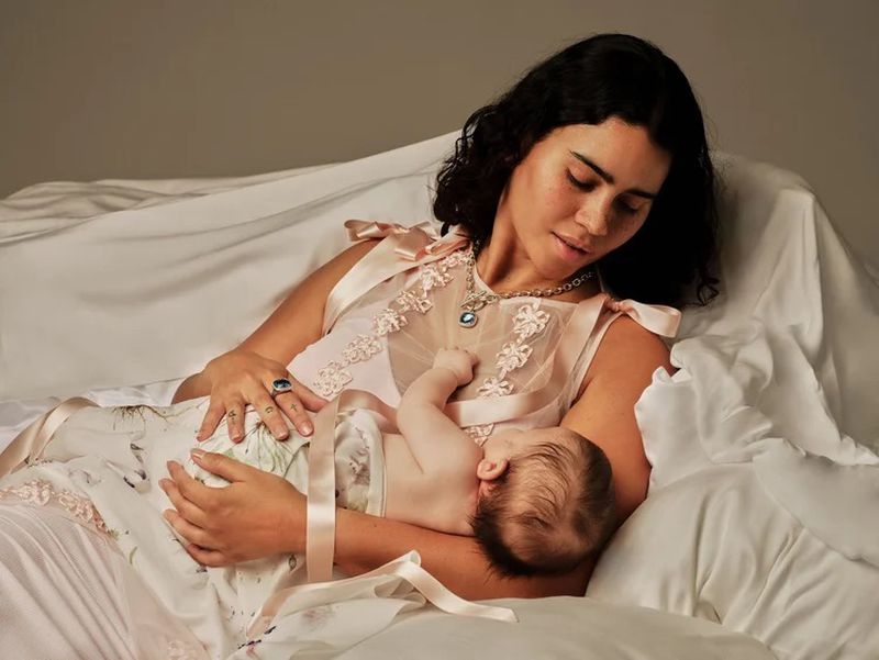 Portrait of a woman in a light pink dress holding a baby in a bright, naturally lit indoor setting with white bedding and soft colors.