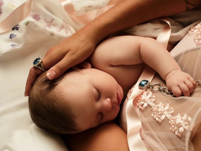 Close-up of a sleeping baby cradled in an adult's arms, adorned with a pink ribbon and a blue gemstone pendant.