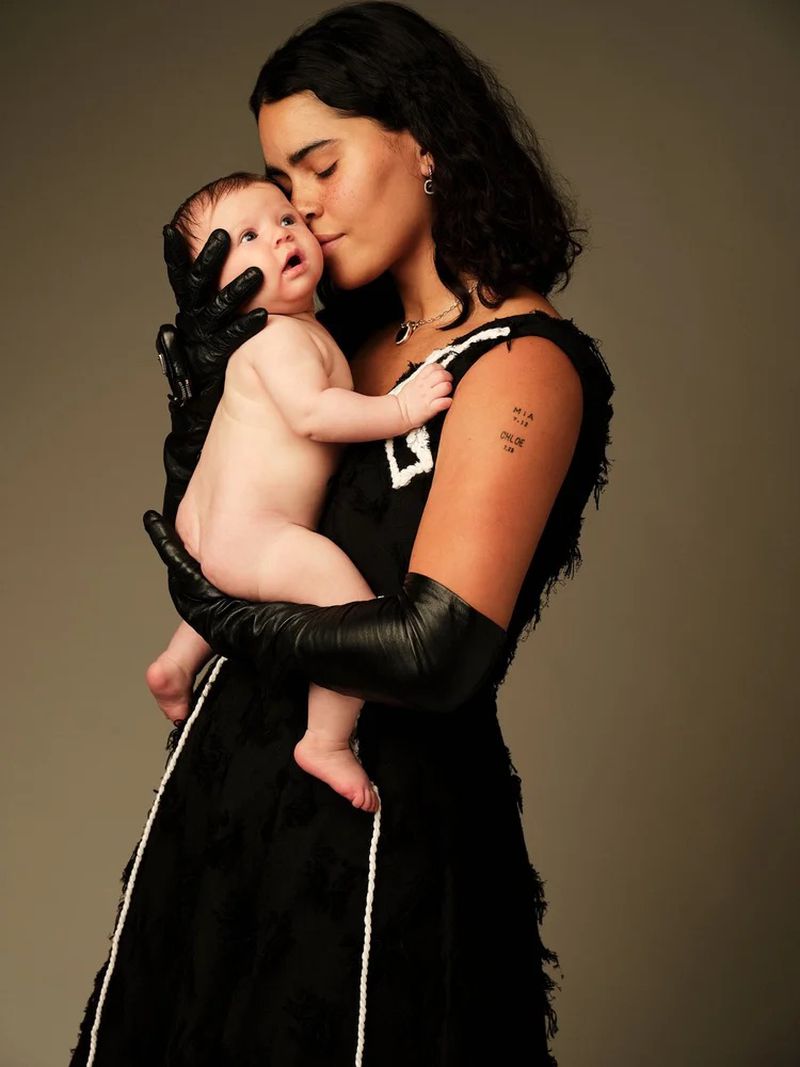 A young woman in a black dress and gloves gently kisses a baby on the forehead, both against a neutral backdrop in a tender moment.
