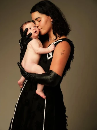 A young woman in a black dress and gloves gently kisses a baby on the forehead, both against a neutral backdrop in a tender moment.