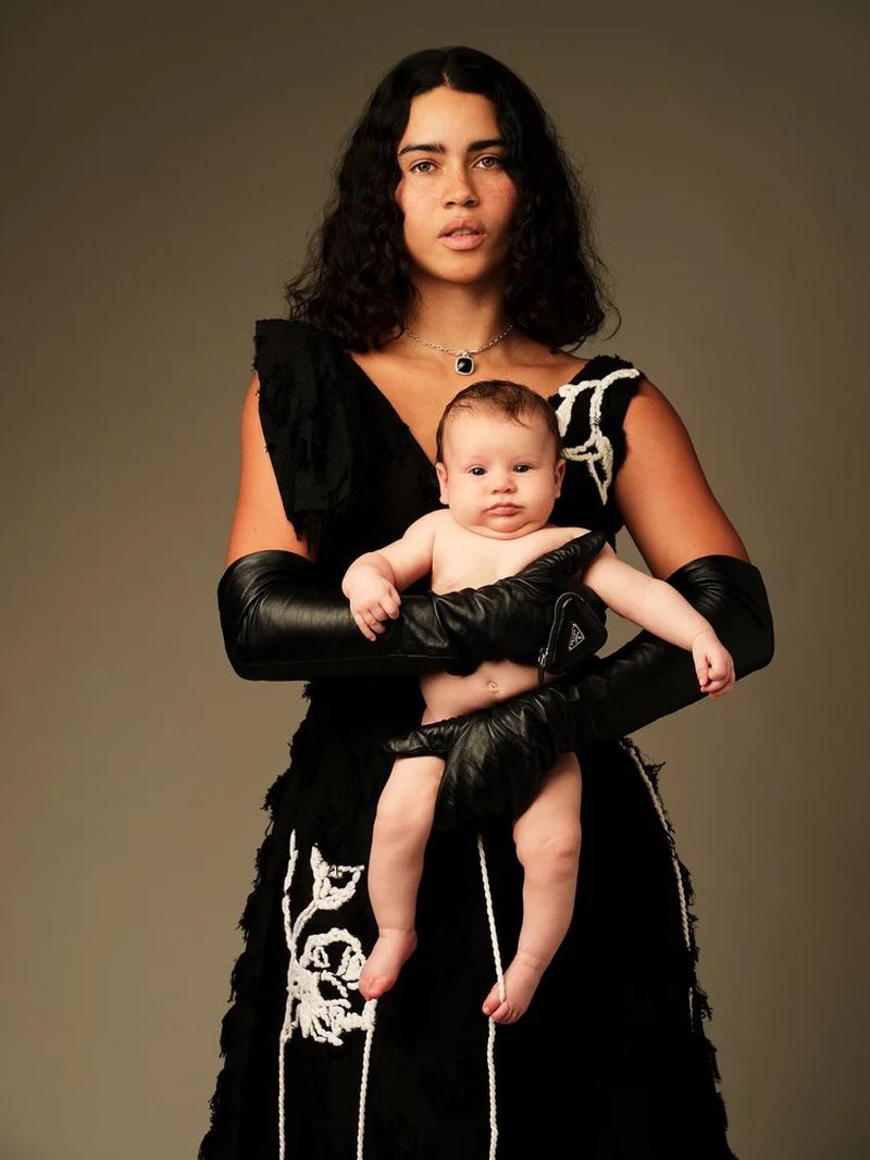 Model Karol Santos wearing a black embroidered dress and long leather gloves while holding an infant against a taupe studio backdrop.