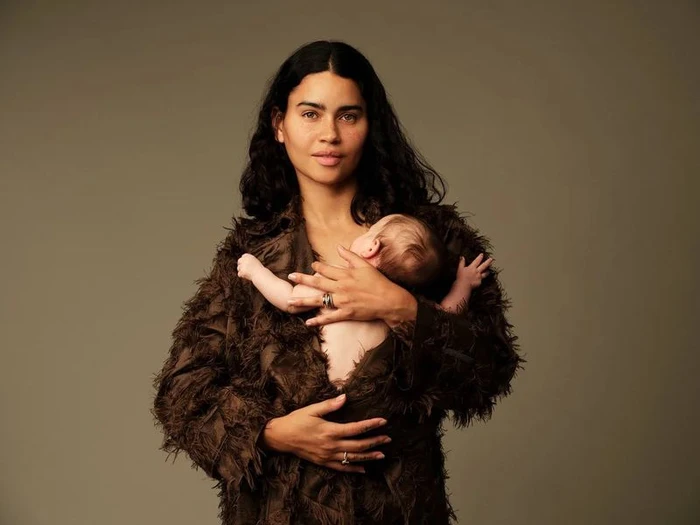 Woman with long dark hair wearing a textured brown coat holding an infant against a neutral grey studio background
