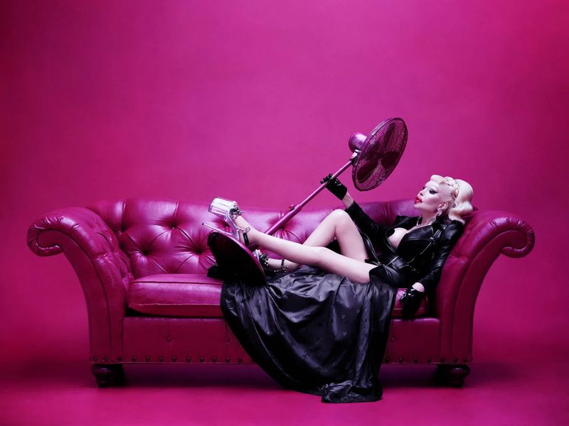 Drag queen reclining on a magenta couch, holding a magenta hand fan in a magenta photo studio with professional lighting.