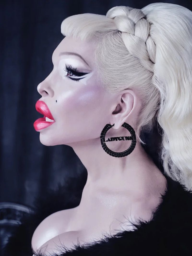Side profile portrait of a person with pale skin, platinum blonde braided hair, heavy makeup, and black hoop earrings with text.