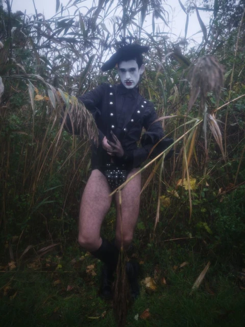 A person in theatrical costume stands in a grassy, reedy field. The dark costume includes a leotard, boots, and stylized hat.