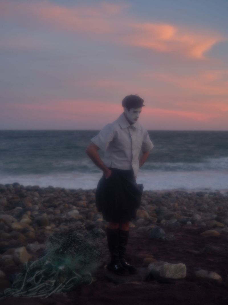 A person stands on a rocky beach near the ocean at sunset wearing a skirt, shirt, and socks.