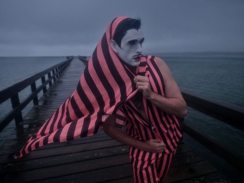 Person in striped suit and face paint poses on a wooden pier with ocean backdrop under an overcast sky, capturing a theatrical moment.
