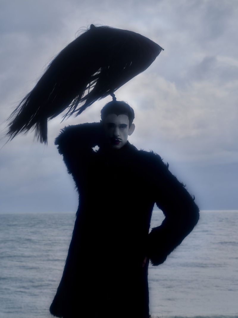 Adult male fashion model poses in a dark coat and unique headdress at the beach under a cloudy sky in a dramatic and surreal fashion image.