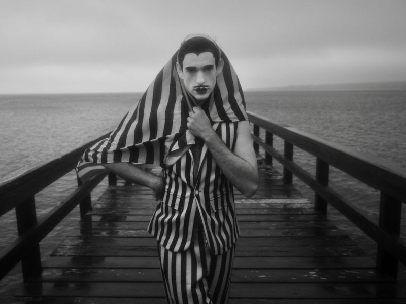 Black and white portrait of a person with striped clothing and makeup, standing on a weathered wooden pier against a calm sea backdrop.