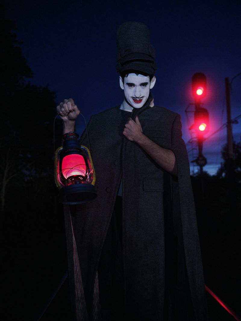 Person in dark coat and hat holds a lantern with red glow at night, standing on railroad tracks with a red traffic light.