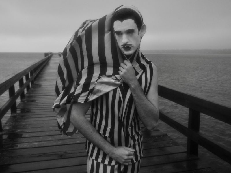 Monochrome portrait of a person in striped clothing on a wooden pier, with dark makeup, wearing a striped cover, seaside backdrop.
