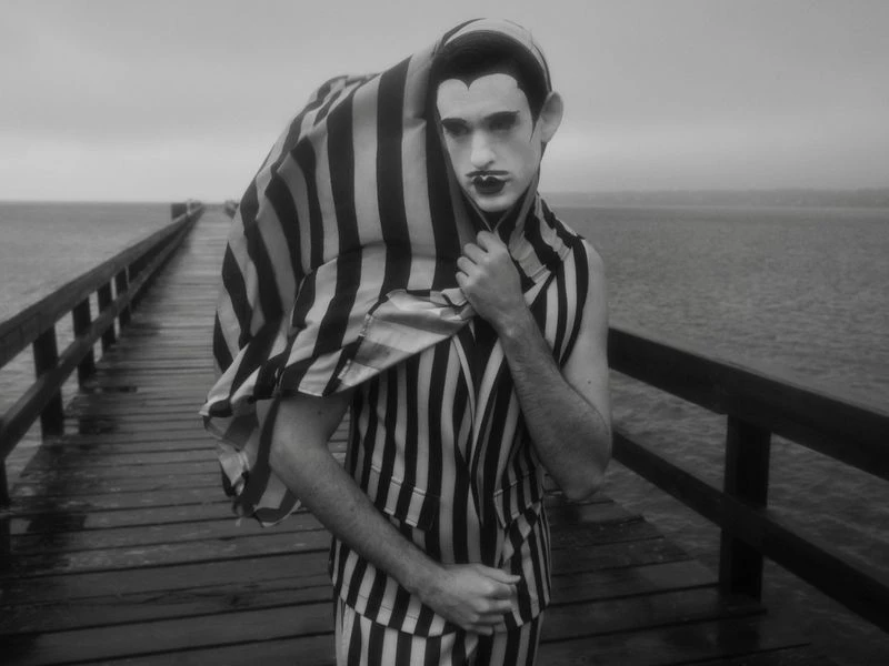 Monochrome portrait of a person in striped clothing on a wooden pier, with dark makeup, wearing a striped cover, seaside backdrop.