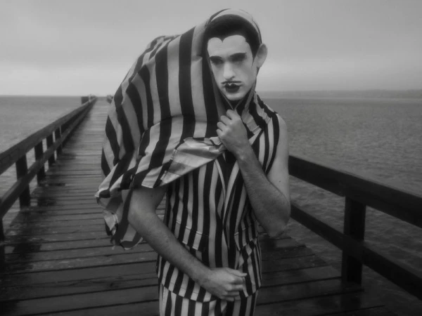 Monochrome portrait of a person in striped clothing on a wooden pier, with dark makeup, wearing a striped cover, seaside backdrop.