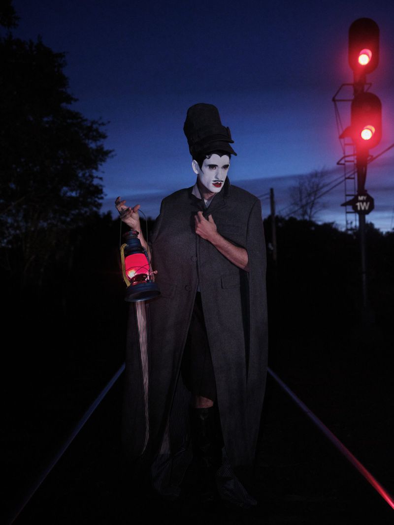 Person in theatrical costume with dark cape and painted face, holding a red lantern on a railroad track at night with a signal light.
