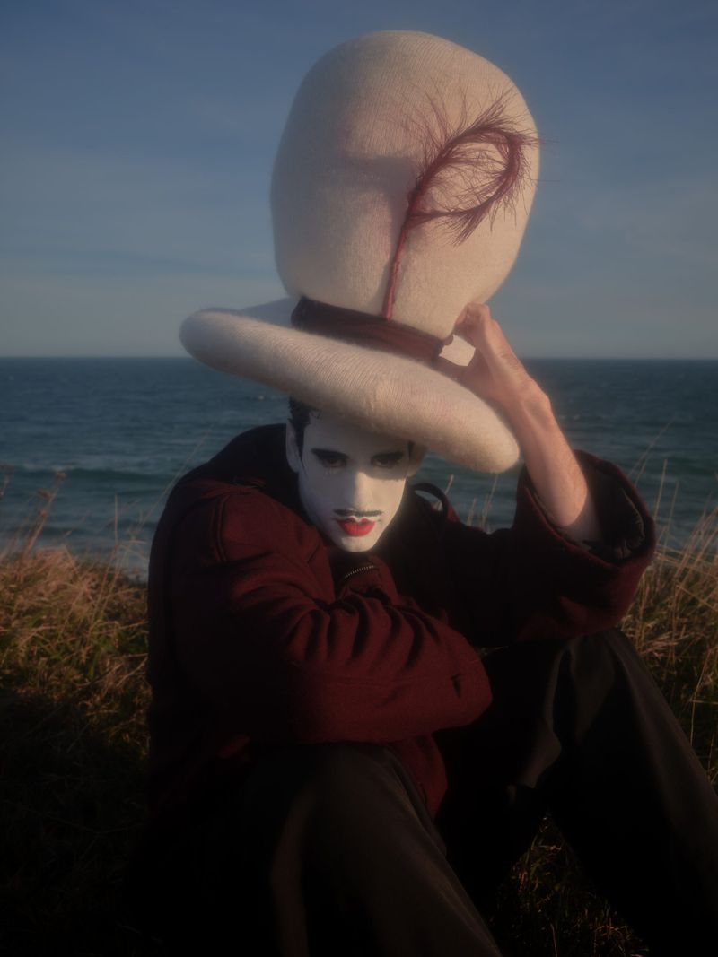 Person in white face paint wears a large hat while seated on grassy land near the ocean under a bright, sunny sky.